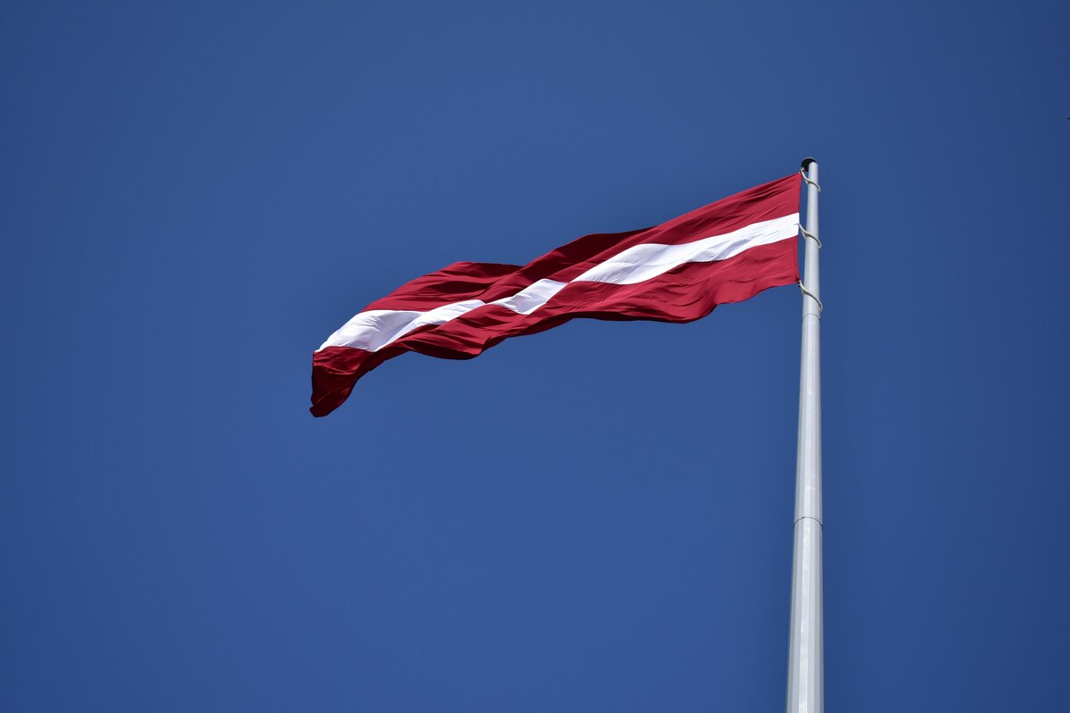 The Latvian flag — carmine red with a white band — flying against a blue sky.