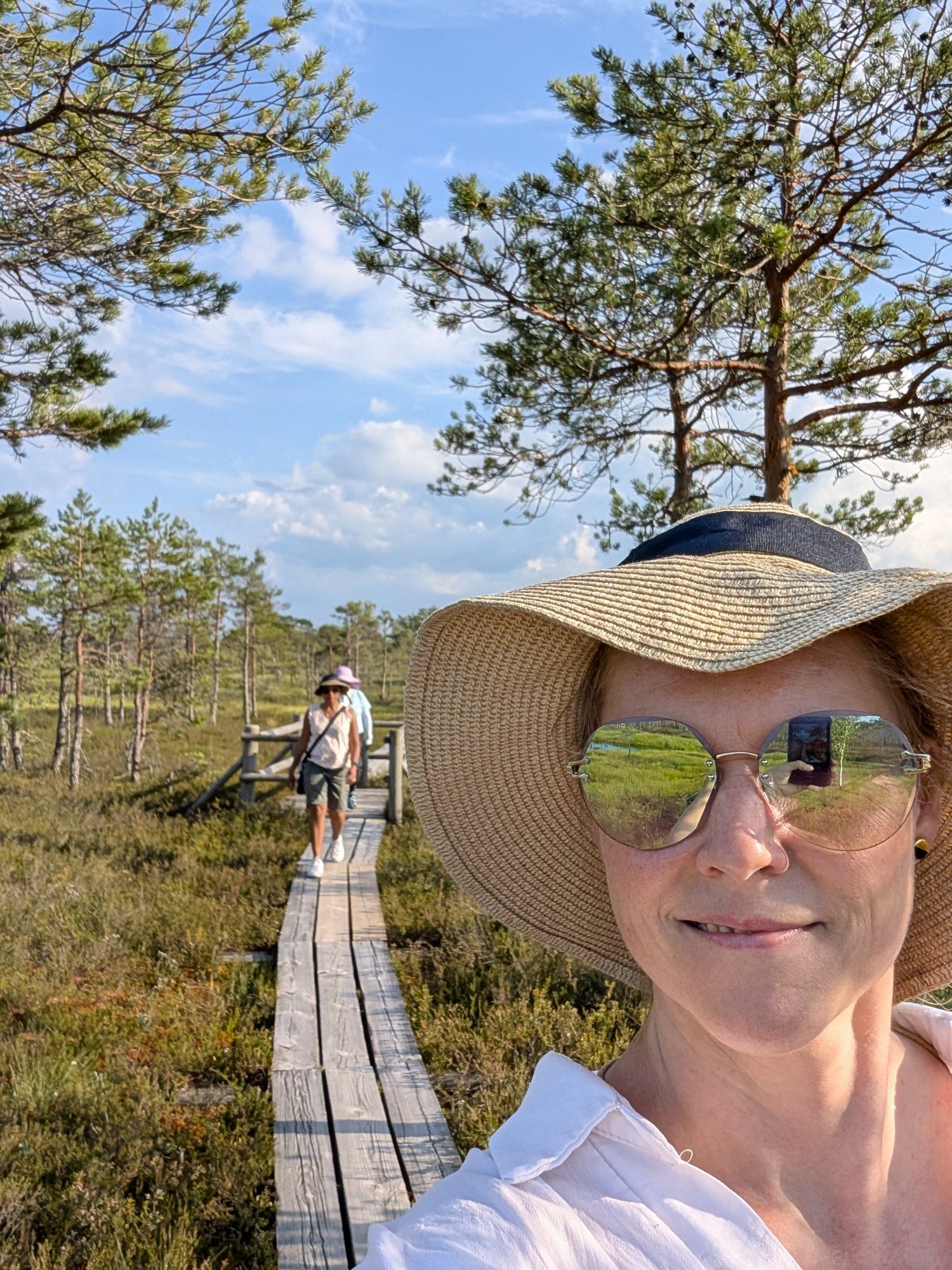 A guest in a wide-brim sun hat on the Ķemeri Bog boardwalk, the raised bog reflected in her sunglasses, with another visitor further along the plank path